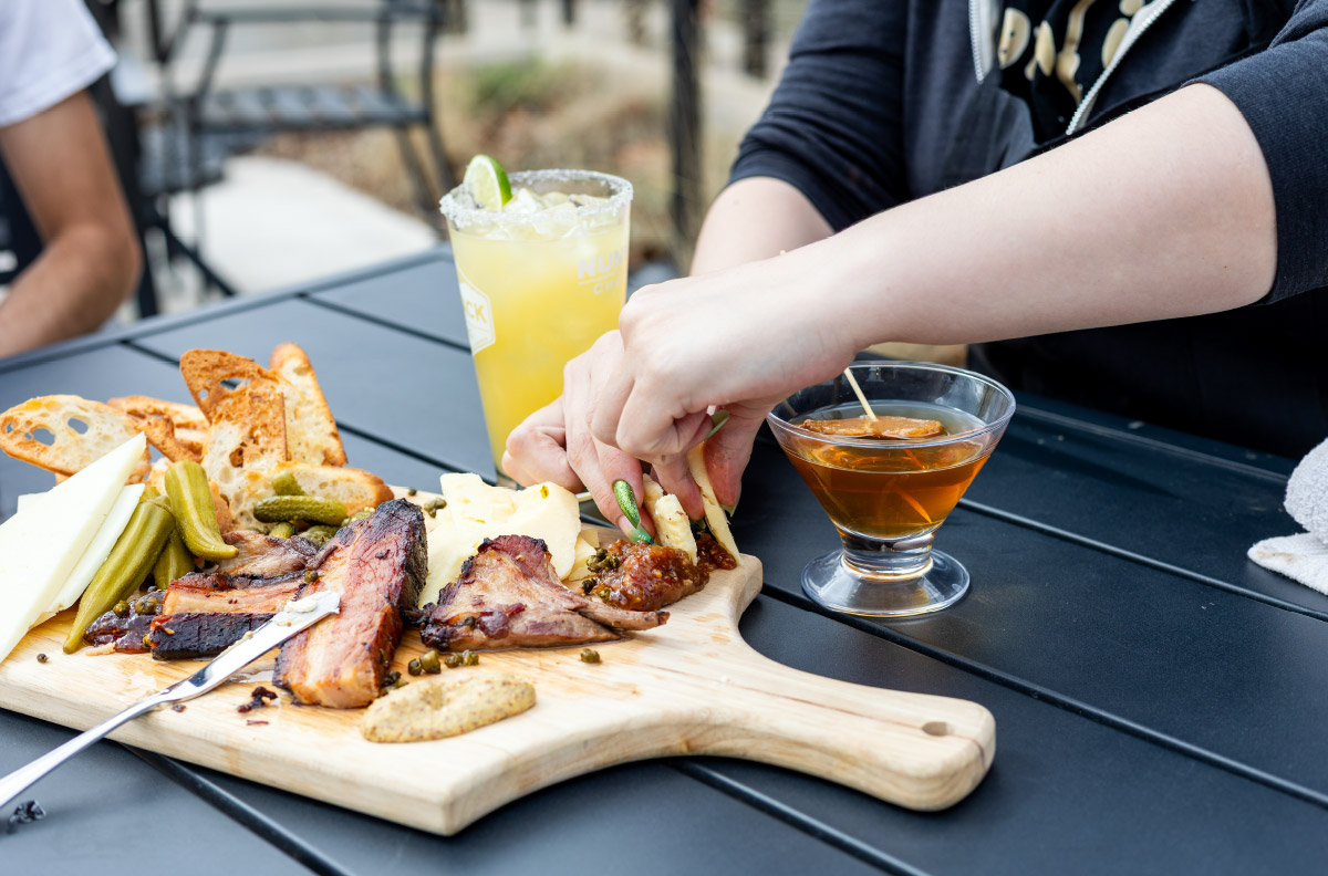 A shared board of smoked meats, cheese and pickles with cocktails on an outdoor patio table